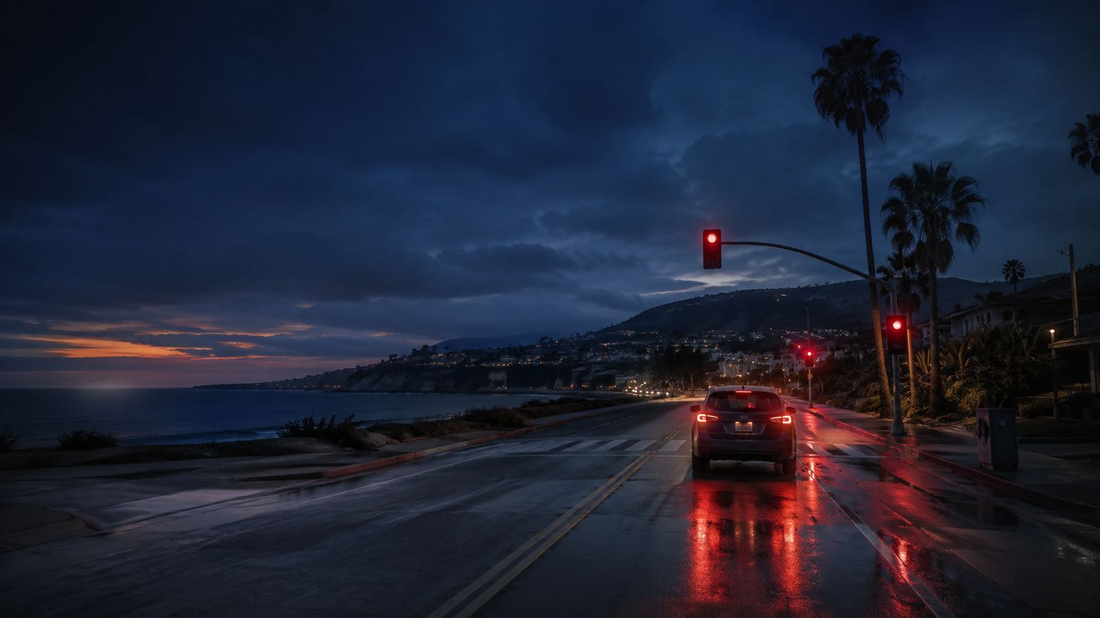 A car stopped at a red light on a wet coastal road at dusk